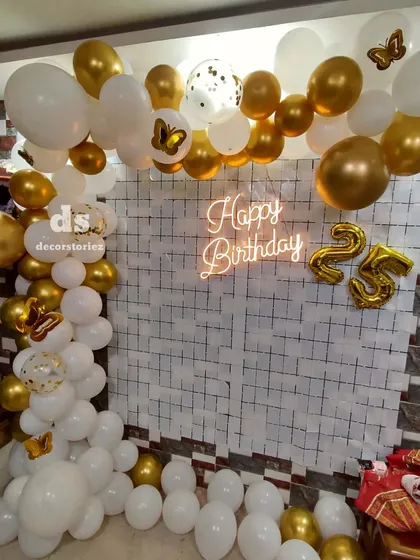 A glamorous white and gold theme for a 25th birthday celebration at home. This decor features a balloon half-arch, a 'Happy Birthday' neon sign, and foil balloons for the age.