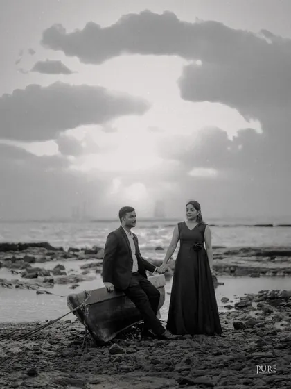 A dramatic black and white photo of Akshay and Kiran on a rocky beach. The cloudy sky and the rustic boat create a moody and cinematic pre-wedding portrait.