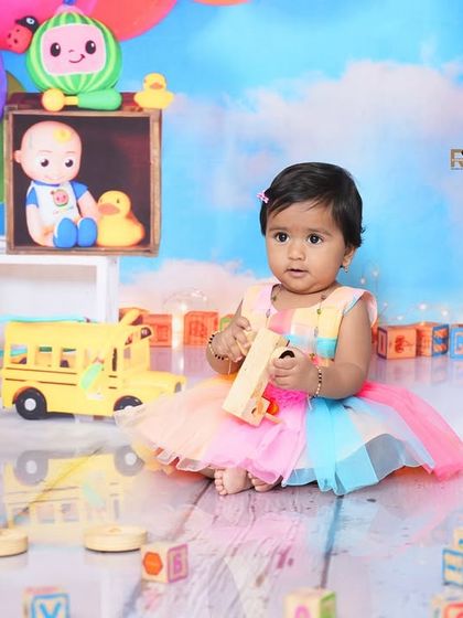 A baby girl in a rainbow dress plays with wooden toys in the Cocomelon-themed studio. The vibrant colors and fun props create a lively and happy atmosphere.