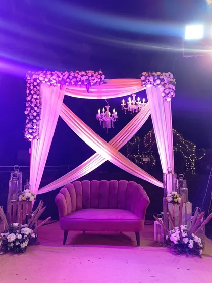 A cozy and chic seating arrangement for a Sangeet or reception. The purple velvet sofa is set against a draped backdrop with a chandelier, creating a perfect spot for photos.
