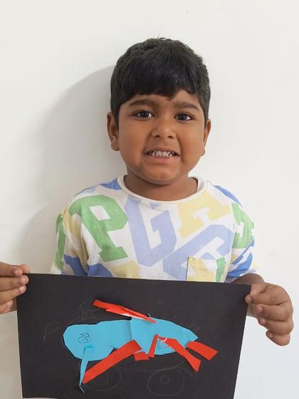 A young boy holds up his paper-craft creation of a crab, made by cutting and pasting colorful shapes.