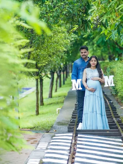 Our outdoor maternity shoots are designed to be a relaxed and enjoyable experience for expecting parents. Here, the couple stands together on a quaint railway track surrounded by lush greenery, holding letters that spell 'MOM'. This playful prop adds a personal touch to their parenthood photoshoot.