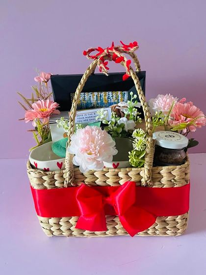 A beautiful couple's hamper in a cane basket. It includes heart-themed mugs, snacks, and floral decorations, perfect for an anniversary or wedding.