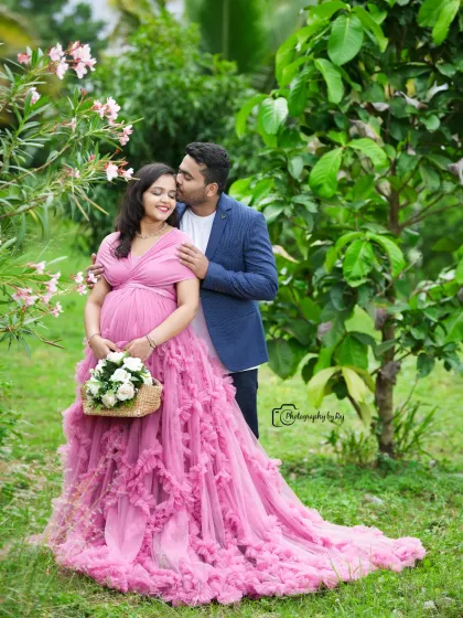 A sweet couple's portrait in a garden, with the father-to-be kissing the mother-to-be's forehead. She is wearing a pink ruffled gown.