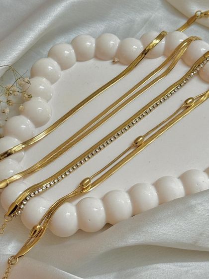 A close-up of delicate gold chain bracelets on a decorative tray. The tight crop and soft lighting emphasize the texture and fine details of each piece.