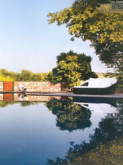 A tranquil moment by the pool at Aishbagh, where the water reflects the surrounding trees and sky. The design of the outdoor space, including the stone wall and comfortable daybed, is focused on creating a peaceful retreat.