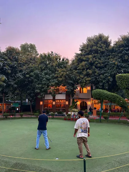 Two friends enjoy a casual putting session at twilight, with the warm lights of the cafe in the background.