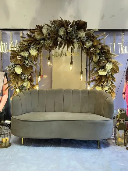A unique boho-chic setup to celebrate a major milestone for two sisters. The dried floral arch, hanging Edison bulbs, and plush velvet sofa create a sophisticated and celebratory stage.