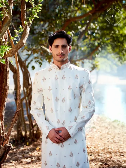 A groom's portrait in a serene outdoor setting, wearing a classic off-white sherwani. The subtle embroidery catches the light beautifully, proving that elegance doesn't need to be loud.