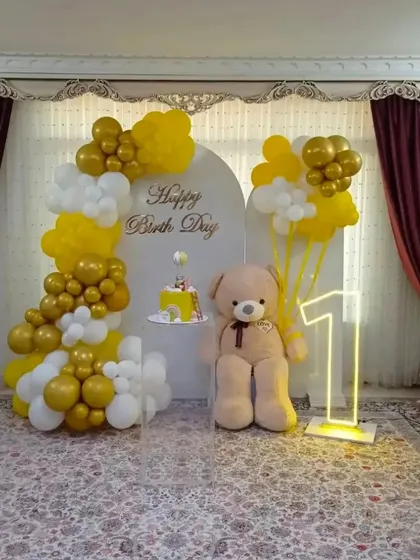 An elegant first birthday setup in yellow and white. This home decoration features a balloon arch against a custom backdrop, a large teddy bear, and a glowing number '1'.