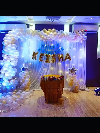 An elegant birthday party setup with a white and gold balloon arch and a sheer curtain backdrop with fairy lights.