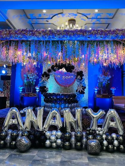 A grand and glamorous setup for Ananya's birthday in a banquet hall. The stage is decorated with a stunning blue and purple lighting scheme, a massive balloon arrangement, and large silver foil letters spelling out her name.