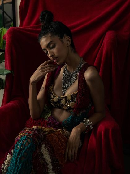 A moody, seated portrait in a rich, red setting. The lighting creates deep shadows that accentuate the textures of the handcrafted garment.