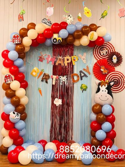A charming farm animal themed birthday party. This decoration includes a balloon arch with cow-print accents, a red fringe curtain, and cute animal cutouts like a cow and a tractor.