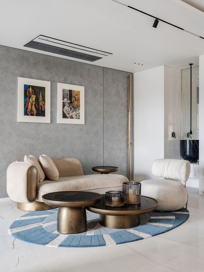 This informal seating area uses a soft, sculptural chaise lounge and nested bronze coffee tables to create a comfortable corner for relaxation. The round, patterned rug defines the space, while the neutral grey wall allows the vibrant artwork to stand out.