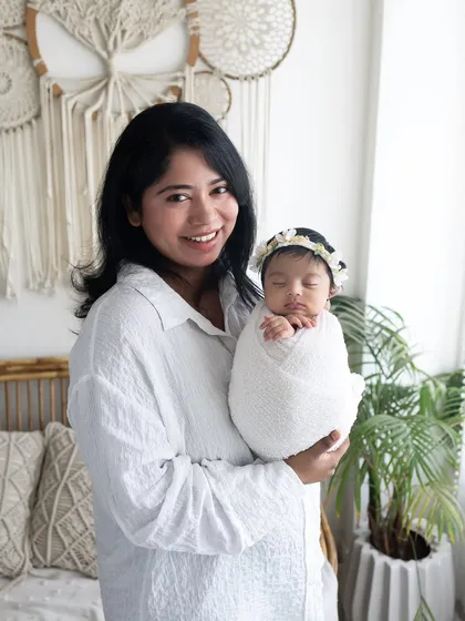 A new mother's joy is radiant in this photo. She is holding her sleeping, swaddled baby against the backdrop of my cozy, boho-inspired studio setup.
