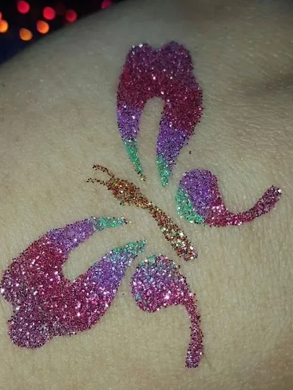 A close-up of an artistic butterfly tattoo. The blend of pink, purple, and gold glitter gives this design a warm, sunset-like feel.