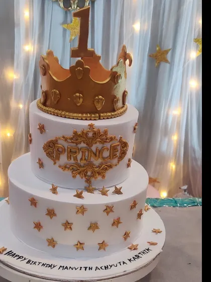 A beautiful three-tiered prince cake in white and gold. The cake is adorned with a large crown, gold stars, and an elegant script, fit for royalty.