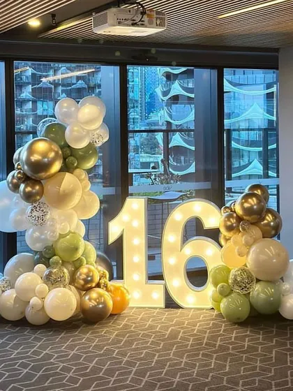 An elegant and simple balloon arc framing "16" marquee numbers. The olive green, gold, and white balloons create a sophisticated color palette for a sweet sixteen party.