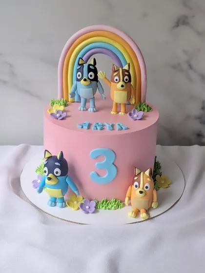 A cheerful cake for a third birthday, featuring Bluey and her family. This pink cake is decorated with a pastel rainbow and cute, handcrafted figures of Bluey and Bingo.