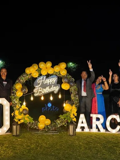 A celebratory group photo from an outdoor 60th birthday party at night, with everyone cheering in front of the large decorative signs.