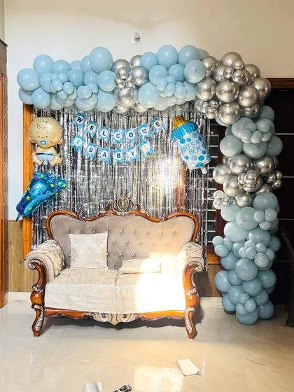 A 'Welcome Baby' setup with a silver tinsel backdrop and an L-shaped balloon arch in shades of blue and silver.