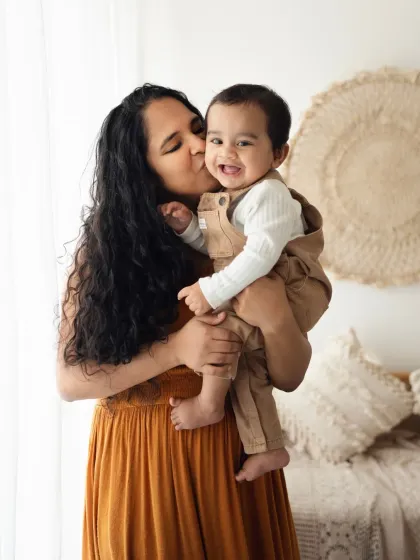 A mother kisses her happy baby's cheek in a warmly lit, boho-style studio. A perfect example of a tender mother and child portrait.
