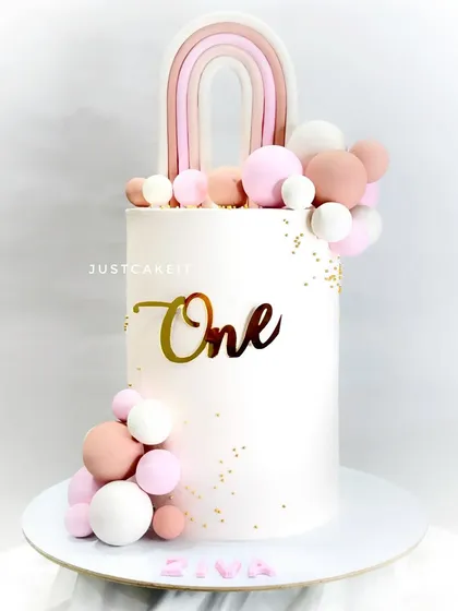 This 10-inch tall cake is a statement piece for a first birthday. The clean white design is accented with a boho rainbow and a cascade of pink and white edible spheres.
