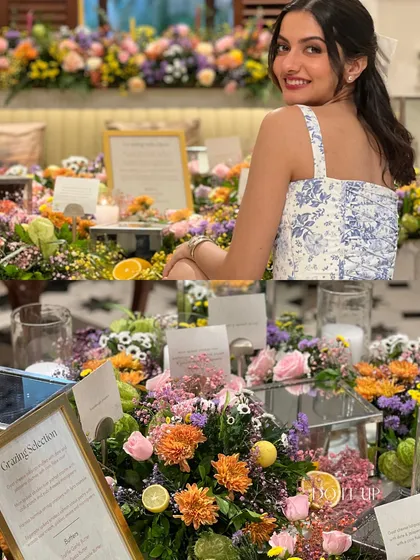 A vibrant grazing table overflowing with colorful flowers. This setup for an intimate gathering shows a woman smiling, surrounded by a beautiful arrangement of florals and personalized details.