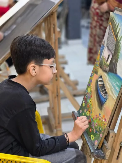 A young student concentrates on his colorful landscape painting, using oil pastels to create a vibrant scene with a boat on a beach.
