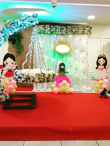 A doll-themed naming ceremony decoration for a baby girl. The stage features a floral arch, a decorated cradle, and cute doll cutouts with pink and yellow balloons.