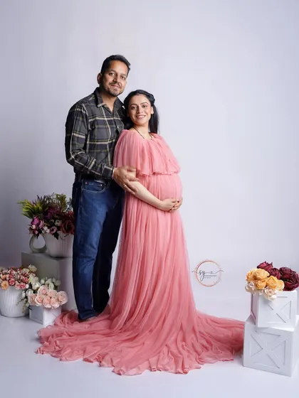 A classic couple's maternity portrait. The expecting mother looks radiant in a flowing pink gown, surrounded by floral arrangements that add a touch of softness and beauty to the studio setting.