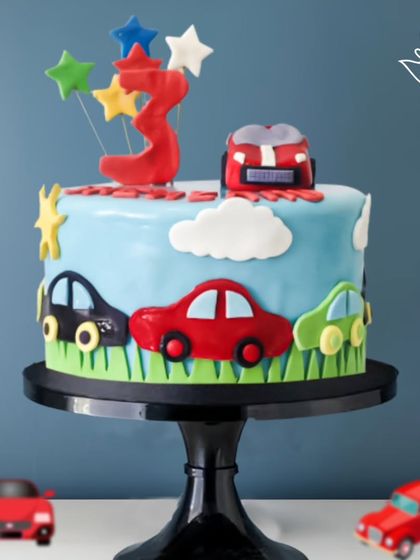 A fun car-themed cake for a 3rd birthday. The design features colorful cars driving on a grassy road under a blue sky, perfect for a little adventurer.