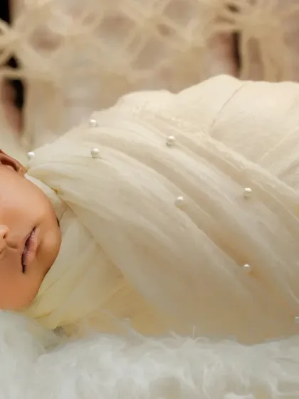 An angelic newborn, wrapped in a pearl-studded cream cloth with a matching flower headband, sleeps on a bed of white fluff.