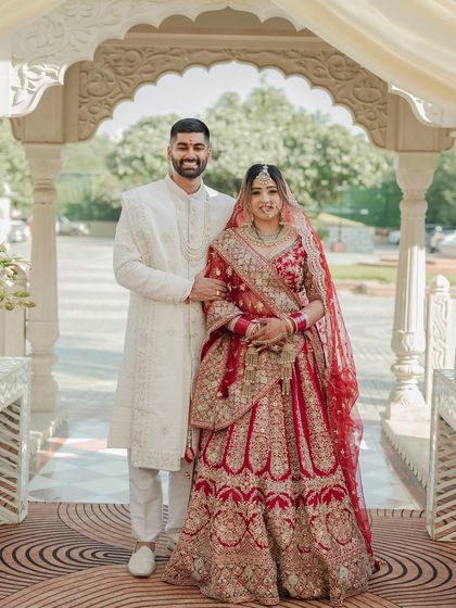 A classic full-length portrait of the bride and groom on their wedding day. Their traditional red and white outfits stand out against the elegant architecture.