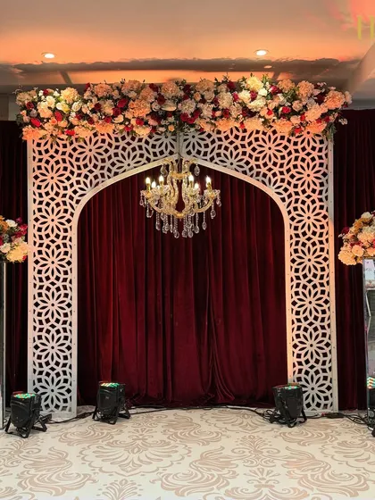 A royal photo booth or small stage setup, featuring a white jali arch against a rich maroon velvet curtain, topped with a floral arrangement and a chandelier.