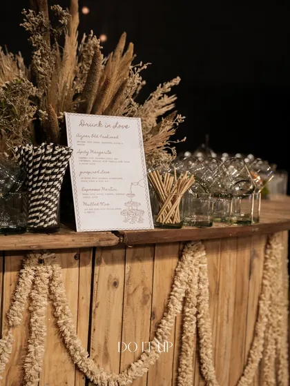 A rustic wooden bar set up for the bohemian Sangeet in Jaipur. It's decorated with garlands of white flowers and dried pampas grass, perfectly matching the earthy theme.