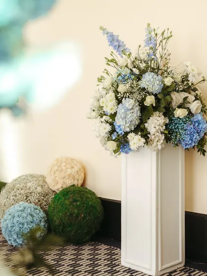 A tall white pedestal with an overflowing arrangement of blue and white hydrangeas, flanked by decorative moss and floral spheres.
