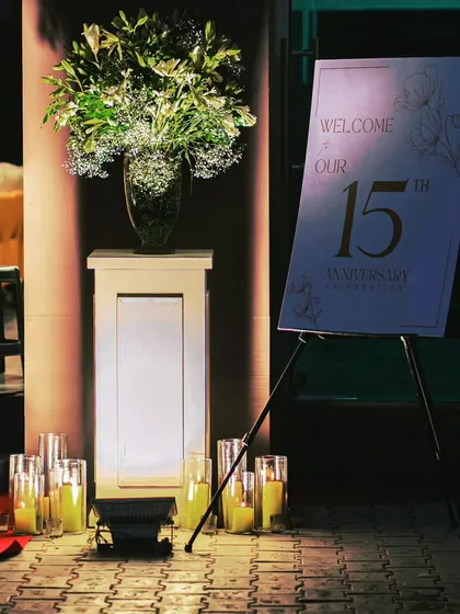 A simple yet elegant welcome corner for a 15th-anniversary event, featuring a floral arrangement, candles, and a custom welcome sign.