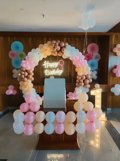 This pastel and rose gold theme is perfect for a stylish first birthday party. The setup includes a half-arch of balloons, a 'Happy Birthday' neon sign, a marquee number '1', and other small balloon details to fill the space.