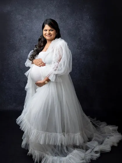 This full-length shot showcases the beautiful design of the white maternity gown. The soft ruffles and long train add a touch of glamour to this elegant studio portrait.