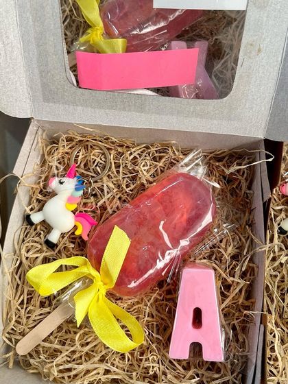 A close-up of a personalized popsicle-themed gift box. This one includes a strawberry popsicle soap, a unicorn keychain, and the initial 'A' in a matching pink soap, all nestled in a charming gift box.