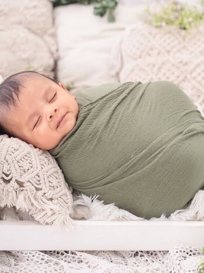A contented smile on a sleeping baby, swaddled in an earthy green wrap and resting on a miniature bed. The neutral tones create a calm and natural feel.