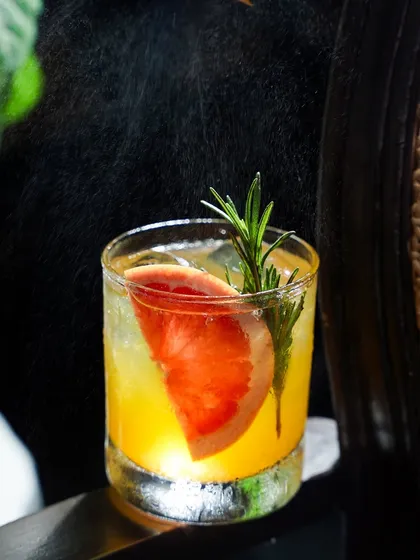 A close-up action shot of a grapefruit and rosemary cocktail being spritzed with zest. The backlighting catches the fine mist, adding a sensory dimension to the image that highlights the freshness of the ingredients.