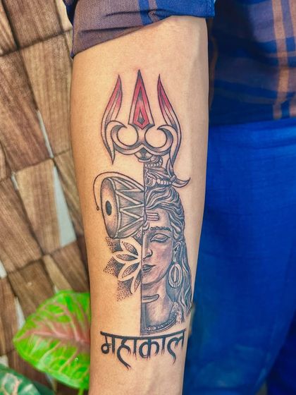 A custom Mahakal tattoo design that combines half of Shiva's face with his Trishul and Damaru. The 'Mahakal' calligraphy at the bottom completes this powerful and artistic piece.