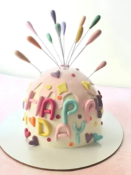 A fun and festive "piñata" style bomb cake. The dome shape is decorated with colorful letters and a firework-like topper, ready for a celebration.
