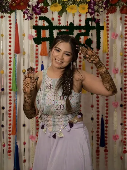 A happy bride poses against a festive "Mehndi" backdrop, her hands held up to display the beautiful artwork we created for her.