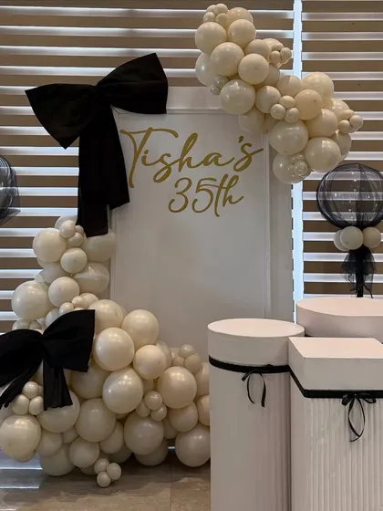 For a chic 35th birthday, we designed a sophisticated black, white, and gold theme. This setup features custom signage, elegant plinths for the cake display, and organic balloon garlands accented with statement bows for a touch of modern glamour.