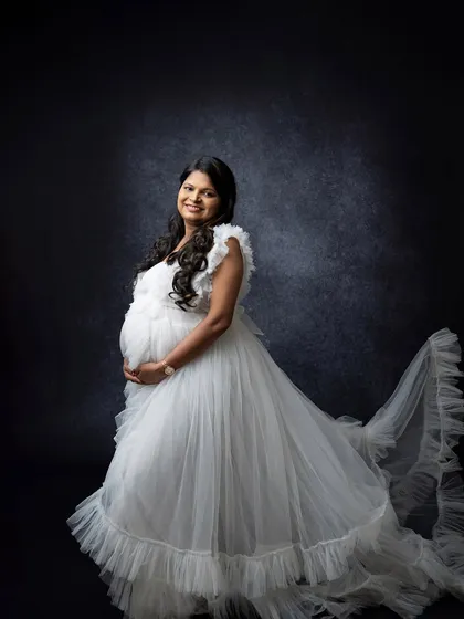 A solo maternity portrait in a flowing white gown against a dark studio background. The contrast creates a timeless and ethereal look, highlighting the mother's joyful expression.