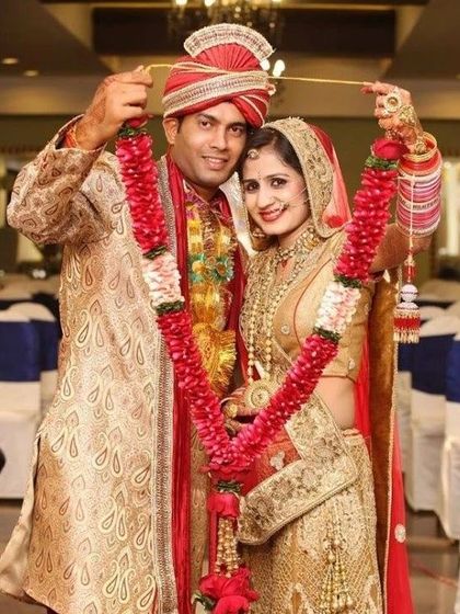 A smiling couple during their varmala ceremony. The groom is wearing a beige paisley-patterned sherwani with a red and gold safa.
